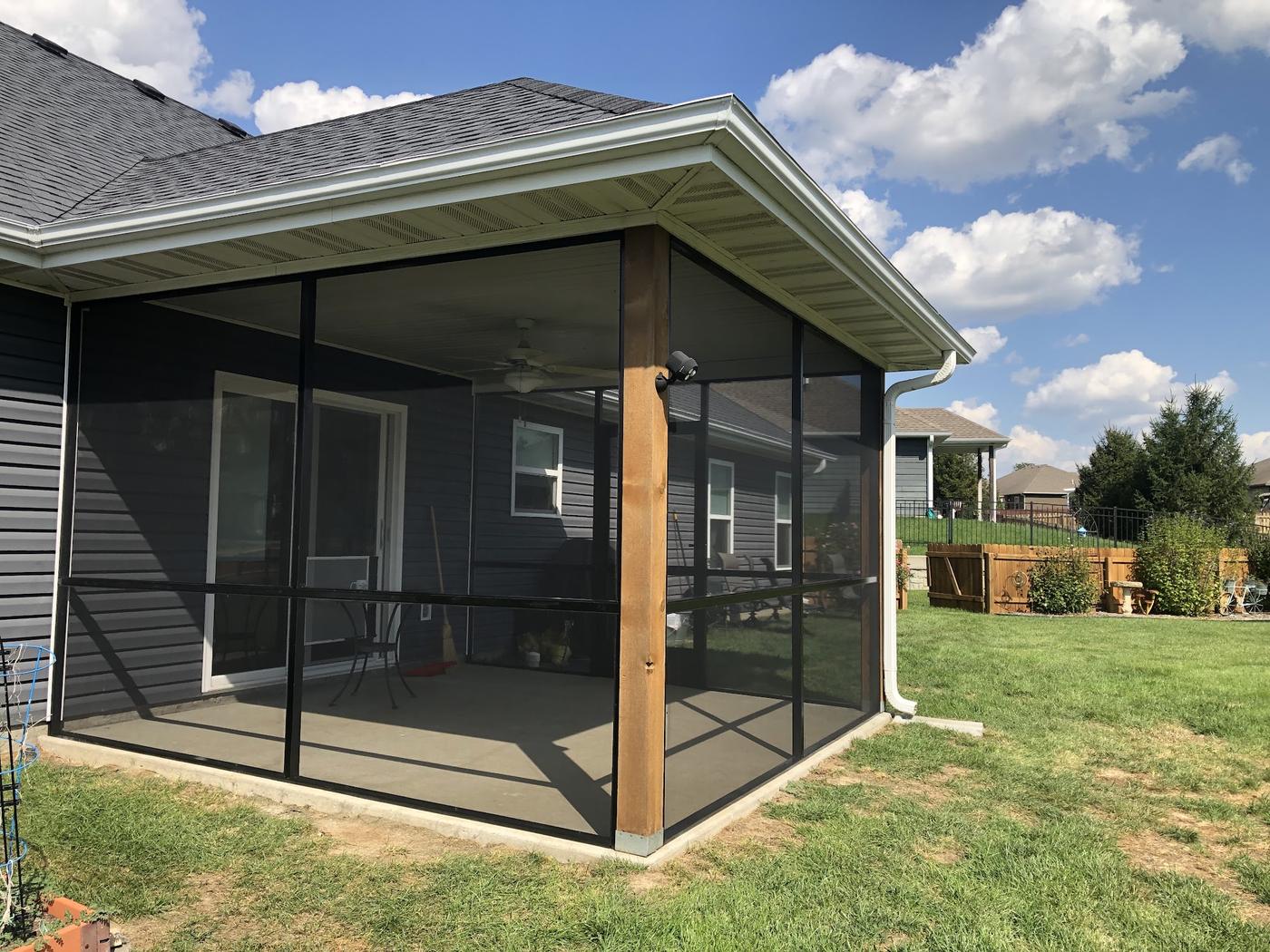 A completed screened porch enclosure under an existing roof, black screen frames around a timber corner post on a concrete slab