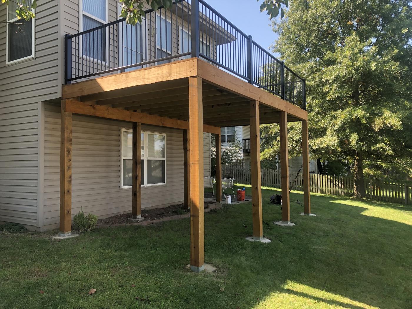 A newly framed elevated deck on heavy timber posts set in concrete footings, beams and joists open underneath, black metal railing above