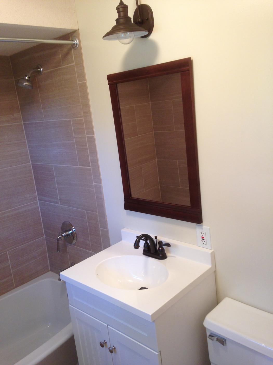 A finished bathroom with a tub surround laid in a running bond, a white vanity, a dark bronze faucet and a framed mirror